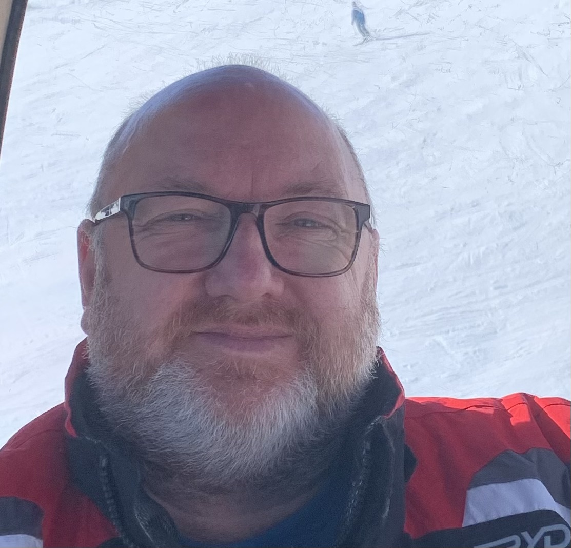 Andy smiling at the camera, wearing black-rimmed glasses, with snow in the background